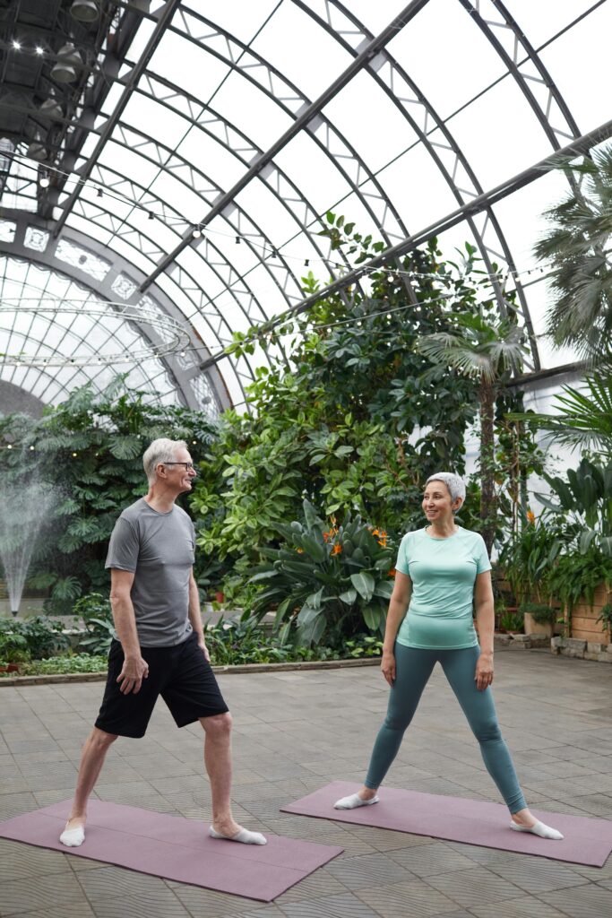 pexels-photo-6787501-6787501 Elderly couple practicing yoga in a lush greenhouse, promoting health and wellness.